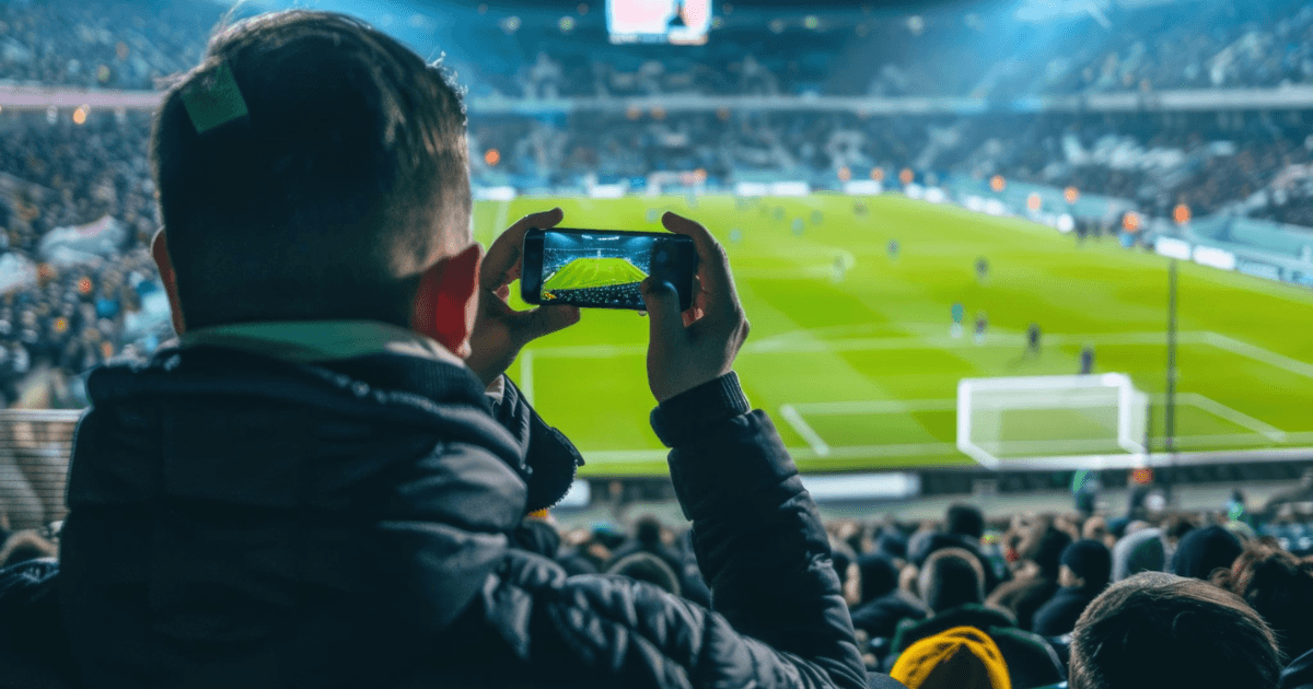 Tilskuer filmer en fodboldkamp med sin mobil fra tribunen på et fyldt stadion